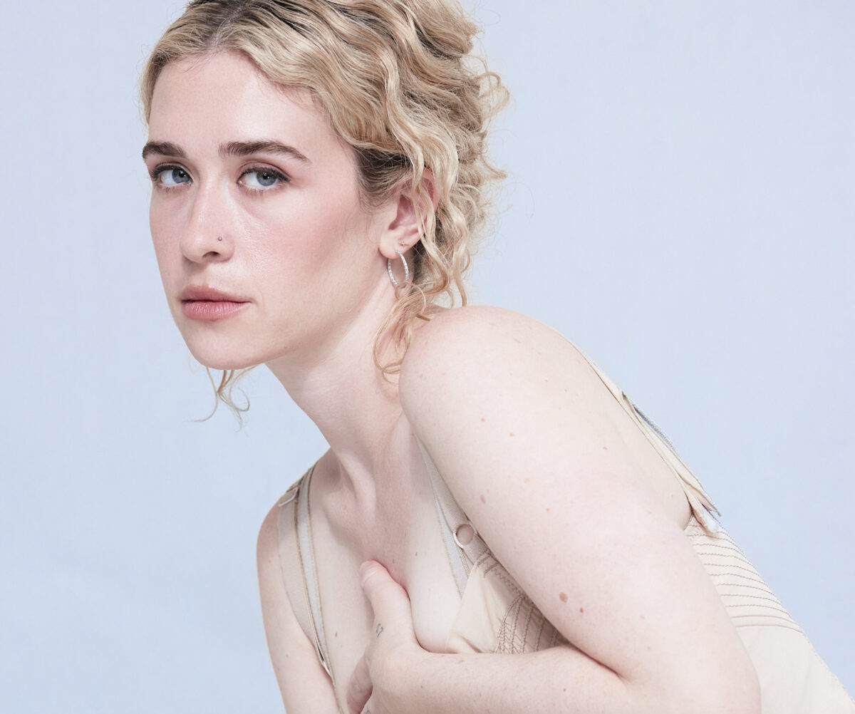 Artist Blondeshell crouching in a nude slip against a light blue backdrop